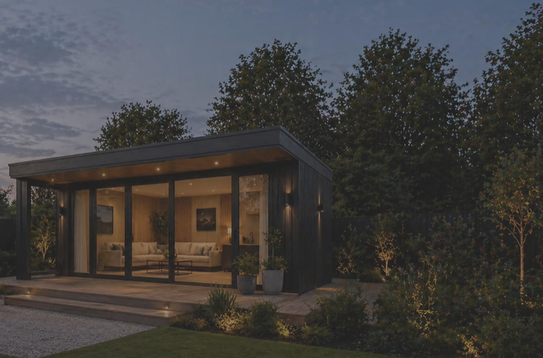 Dark-clad garden studio at dusk with warm interior lighting, a glazed front and a landscaped gravel path